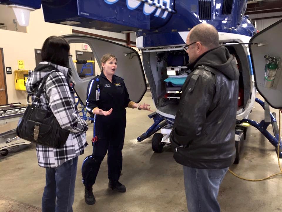 Kristen Cline, RN, in a flight suit beside an air-medical helicopter, walking two people through the aircraft's patient-care configuration